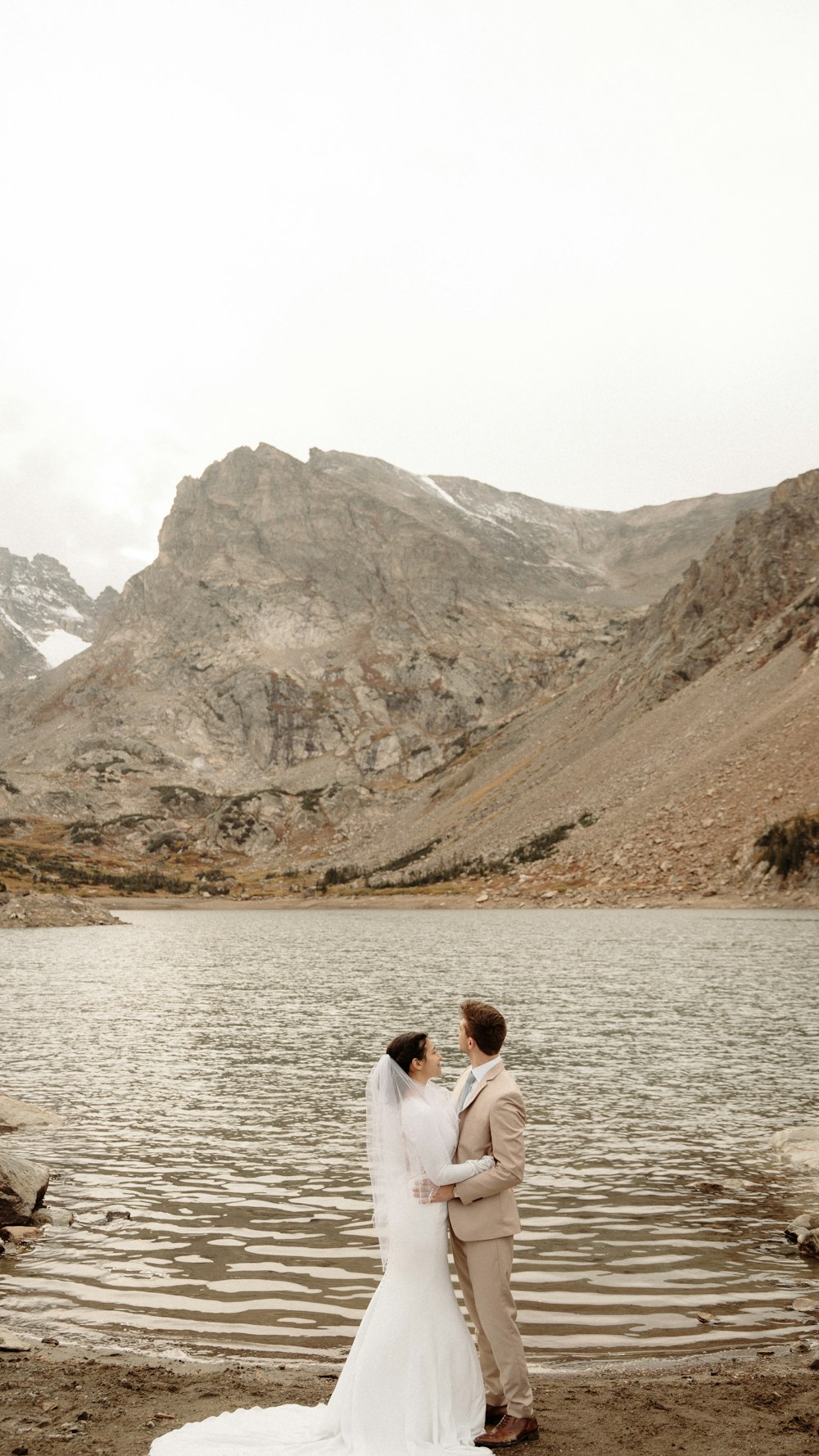 a bride and groom standing in front of a mountain lake