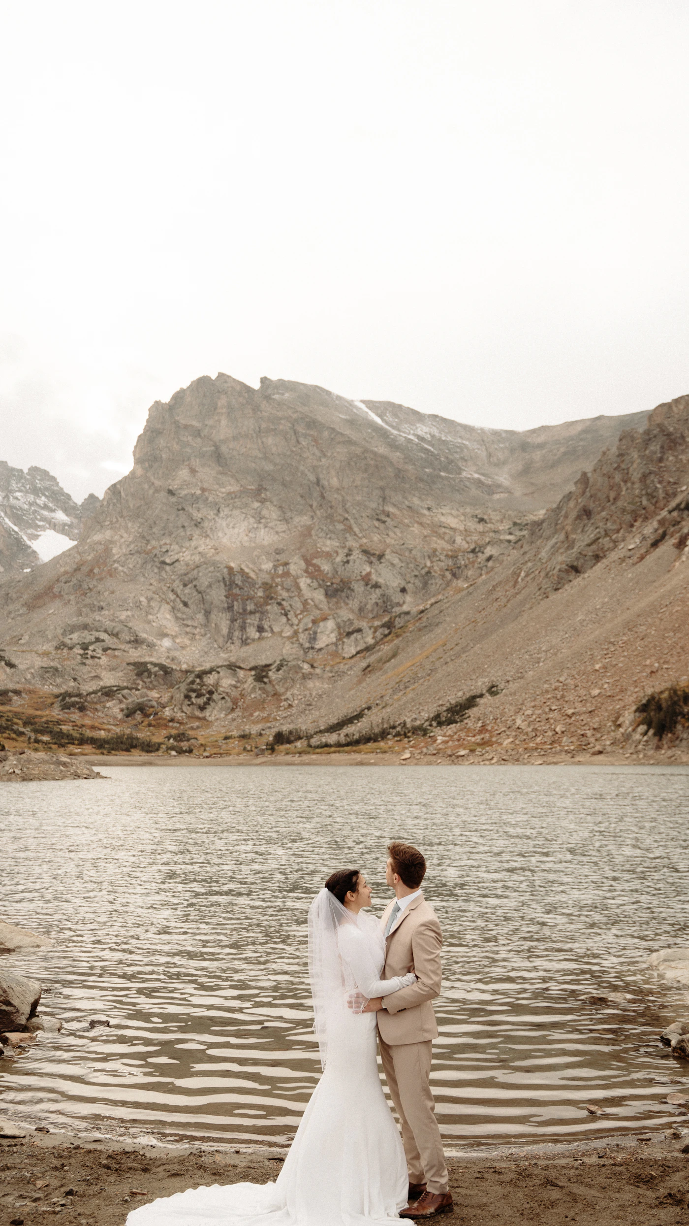 Couple in winter elopement attire standing on the Abraham Lake ice with the frozen bubble formations creating a patterned surface beneath them and the vast Bighorn Wildland mountain landscape extending to the horizon in all directions
