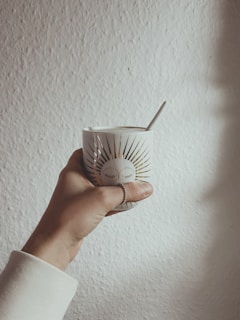 A hand is holding a white mug with a sunburst design and a face with closed eyes printed on it. The mug is held against a textured cream-colored wall. A spoon is resting inside the mug, and the hand is adorned with a ring.