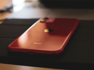 A red smartphone lies on a dark surface, featuring a glossy finish and a prominent logo in the center. The lighting highlights the sleek design and vibrant color of the device.