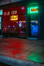 A storefront featuring a nail spa and Thai bodywork shop is illuminated by vibrant neon signs. The red neon sign for the nail spa is prominently displayed on the left, while the right side has a green and blue neon sign for Thai bodywork. Reflections of city lights can be seen on the wet pavement in front.