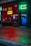 A storefront featuring a nail spa and Thai bodywork shop is illuminated by vibrant neon signs. The red neon sign for the nail spa is prominently displayed on the left, while the right side has a green and blue neon sign for Thai bodywork. Reflections of city lights can be seen on the wet pavement in front.