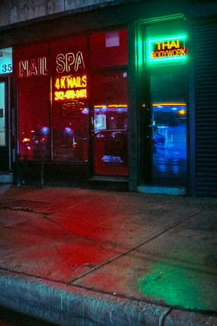 A storefront featuring a nail spa and Thai bodywork shop is illuminated by vibrant neon signs. The red neon sign for the nail spa is prominently displayed on the left, while the right side has a green and blue neon sign for Thai bodywork. Reflections of city lights can be seen on the wet pavement in front.