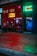 A storefront featuring a nail spa and Thai bodywork shop is illuminated by vibrant neon signs. The red neon sign for the nail spa is prominently displayed on the left, while the right side has a green and blue neon sign for Thai bodywork. Reflections of city lights can be seen on the wet pavement in front.