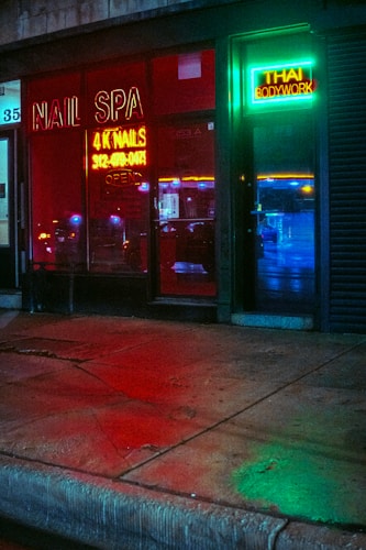 A storefront featuring a nail spa and Thai bodywork shop is illuminated by vibrant neon signs. The red neon sign for the nail spa is prominently displayed on the left, while the right side has a green and blue neon sign for Thai bodywork. Reflections of city lights can be seen on the wet pavement in front.