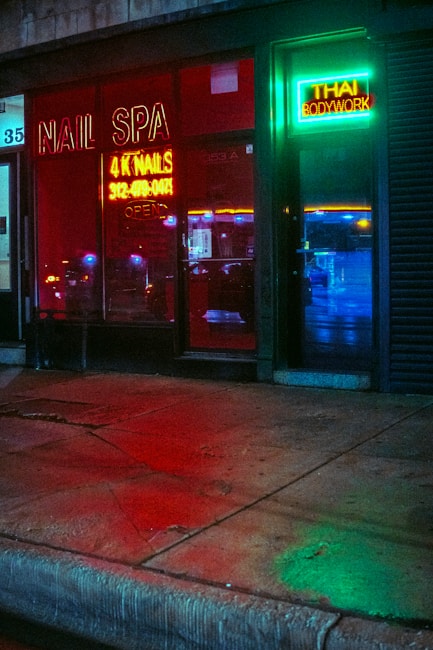 A storefront featuring a nail spa and Thai bodywork shop is illuminated by vibrant neon signs. The red neon sign for the nail spa is prominently displayed on the left, while the right side has a green and blue neon sign for Thai bodywork. Reflections of city lights can be seen on the wet pavement in front.