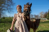 A young girl wearing a floral dress and a headband stands next to a brown pony, both in a grassy field on a clear day. The girl is smiling, holding a metal bucket in her left hand while resting her right hand on the pony. Another smaller pony is in the background. The sky is bright blue with scattered trees and a fenced area behind them.