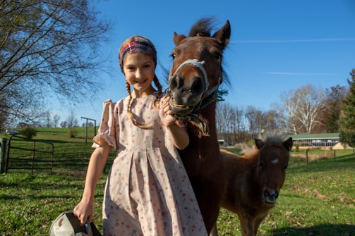 A young girl wearing a floral dress and a headband stands next to a brown pony, both in a grassy field on a clear day. The girl is smiling, holding a metal bucket in her left hand while resting her right hand on the pony. Another smaller pony is in the background. The sky is bright blue with scattered trees and a fenced area behind them.