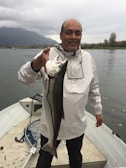 A person is standing on a boat holding a large fish. The background shows a calm body of water with mountains shrouded in clouds. The person is wearing a light-colored rain jacket.