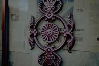 A decorative metal ornament with an intricate floral and leaf design is affixed to a glass surface. The background consists of old newspaper pages with text and images, printed in a foreign language.