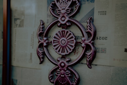 A decorative metal ornament with an intricate floral and leaf design is affixed to a glass surface. The background consists of old newspaper pages with text and images, printed in a foreign language.