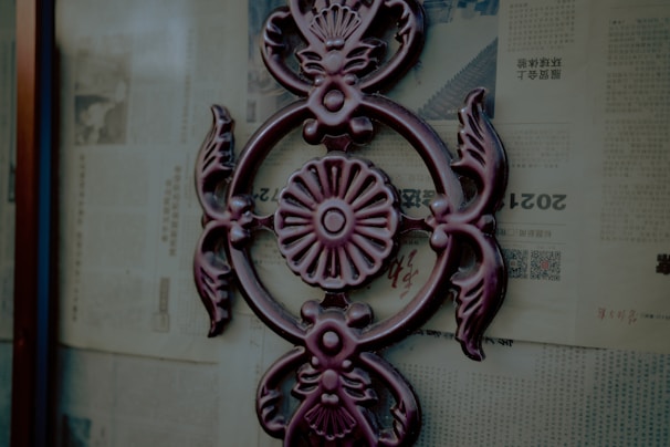 A decorative metal ornament with an intricate floral and leaf design is affixed to a glass surface. The background consists of old newspaper pages with text and images, printed in a foreign language.