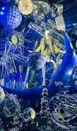 A lavish display combines celestial and astronomical themes, featuring a mannequin wearing a gold sequin outfit seated on a blue crescent moon. Surrounding elements include a disco ball, intricate geometric structures, a clock with a starburst motif, and various reflective and metallic objects. The scene is richly textured with a space-themed backdrop, highlighted by vibrant lighting.