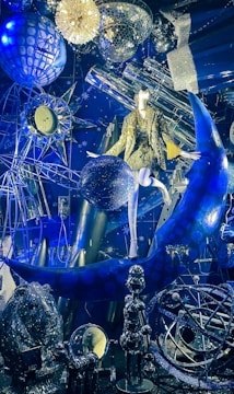 A lavish display combines celestial and astronomical themes, featuring a mannequin wearing a gold sequin outfit seated on a blue crescent moon. Surrounding elements include a disco ball, intricate geometric structures, a clock with a starburst motif, and various reflective and metallic objects. The scene is richly textured with a space-themed backdrop, highlighted by vibrant lighting.