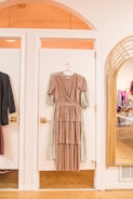 A long, layered brown dress on a hanger is displayed against a white door in a stylish boutique setting. To the right, a large mirror reflects the interior of the store, showing racks of clothes. Warm lighting enhances the cozy, inviting atmosphere.