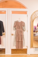A long, layered brown dress on a hanger is displayed against a white door in a stylish boutique setting. To the right, a large mirror reflects the interior of the store, showing racks of clothes. Warm lighting enhances the cozy, inviting atmosphere.