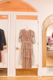 A long, layered brown dress on a hanger is displayed against a white door in a stylish boutique setting. To the right, a large mirror reflects the interior of the store, showing racks of clothes. Warm lighting enhances the cozy, inviting atmosphere.