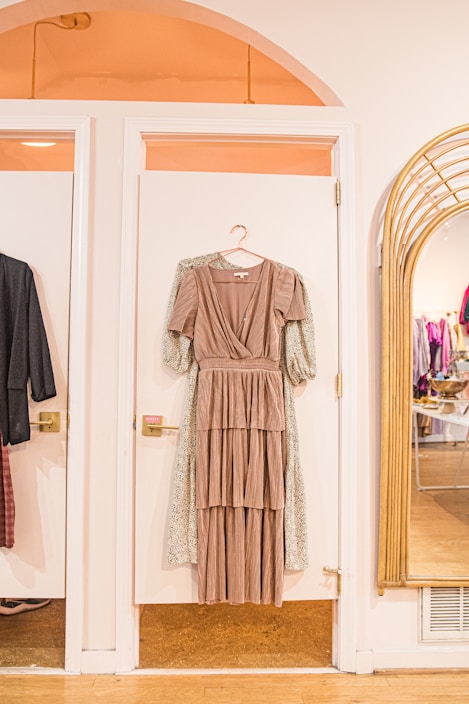 A long, layered brown dress on a hanger is displayed against a white door in a stylish boutique setting. To the right, a large mirror reflects the interior of the store, showing racks of clothes. Warm lighting enhances the cozy, inviting atmosphere.