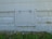 A white, rectangular metal access panel is mounted on the lower part of an exterior wall. The wall has a textured pattern resembling bricks and exhibits patches of peeling paint. Below the panel, there is a patch of overgrown grass and small yellow wildflowers.
