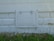 A white, rectangular metal access panel is mounted on the lower part of an exterior wall. The wall has a textured pattern resembling bricks and exhibits patches of peeling paint. Below the panel, there is a patch of overgrown grass and small yellow wildflowers.