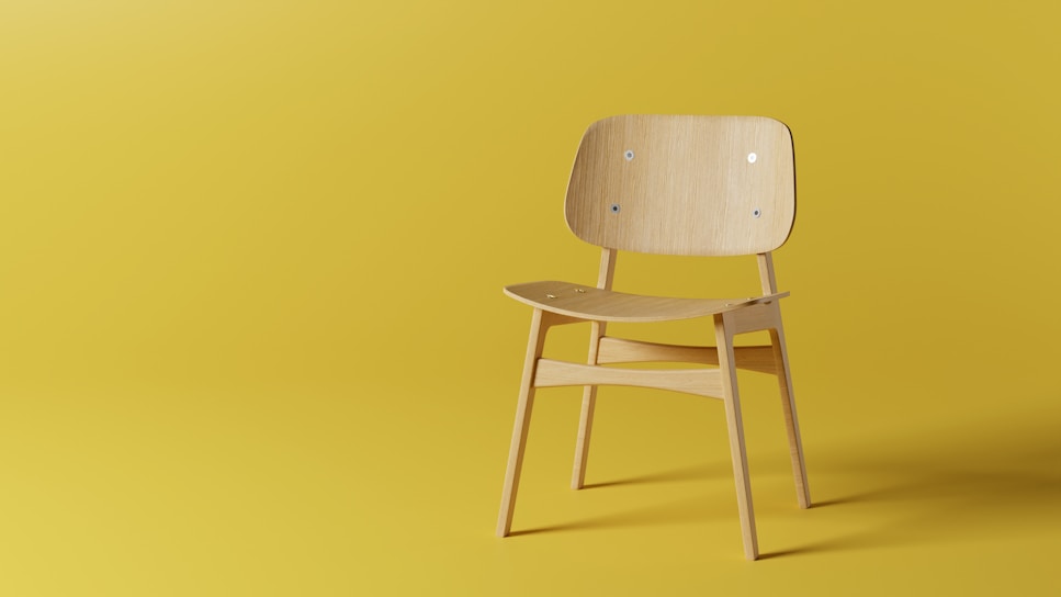 A minimalist wooden chair with a curved backrest and seat is positioned against a solid yellow background. The chair features simple, clean lines and a natural wood finish, contributing to a modern aesthetic.