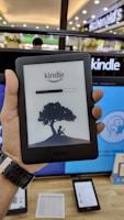 A person is holding an Amazon Kindle e-reader featuring a loading screen with an illustration of a person reading under a tree. In the background, there are several electronic devices displayed on a counter and blurred signage from a store.