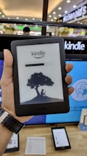 A person is holding an Amazon Kindle e-reader featuring a loading screen with an illustration of a person reading under a tree. In the background, there are several electronic devices displayed on a counter and blurred signage from a store.