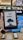 A person is holding an Amazon Kindle e-reader featuring a loading screen with an illustration of a person reading under a tree. In the background, there are several electronic devices displayed on a counter and blurred signage from a store.