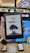 A person is holding an Amazon Kindle e-reader featuring a loading screen with an illustration of a person reading under a tree. In the background, there are several electronic devices displayed on a counter and blurred signage from a store.