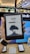 A person is holding an Amazon Kindle e-reader featuring a loading screen with an illustration of a person reading under a tree. In the background, there are several electronic devices displayed on a counter and blurred signage from a store.