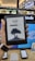 A person is holding an Amazon Kindle e-reader featuring a loading screen with an illustration of a person reading under a tree. In the background, there are several electronic devices displayed on a counter and blurred signage from a store.