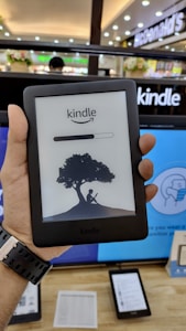 A person is holding an Amazon Kindle e-reader featuring a loading screen with an illustration of a person reading under a tree. In the background, there are several electronic devices displayed on a counter and blurred signage from a store.