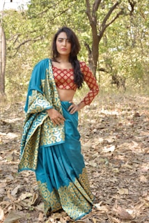 A woman stands in a forest setting, wearing a traditional Indian sari with a blue base and intricate gold patterns. Her blouse is red with a geometric design. The background consists of trees with green foliage and a ground covered with dry leaves.
