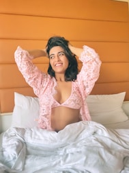 A woman is sitting up in bed, wearing a pink lace robe with matching undergarments. Her arms are raised, hands in her hair, and she is smiling. The bed has white sheets and pillows, and the headboard is orange.
