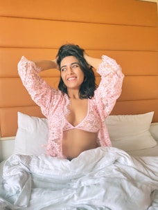 A woman is sitting up in bed, wearing a pink lace robe with matching undergarments. Her arms are raised, hands in her hair, and she is smiling. The bed has white sheets and pillows, and the headboard is orange.