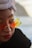 A close-up portrait of a person wearing butterfly-shaped sunglasses with orange and yellow lenses, a white turban-style headwrap, red lipstick, and a black fur-like coat. The background appears to be an out-of-focus coastal or mountainous area.