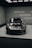 A sleek, dark-colored sports car, specifically a Porsche 911, is displayed indoors on a platform. The setting is modern, with a grid-like ceiling and minimalist walls. Fluorescent lights illuminate the car, highlighting its glossy finish and aerodynamic design.