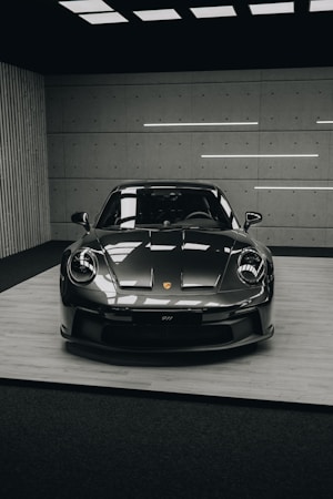 A sleek, dark-colored sports car, specifically a Porsche 911, is displayed indoors on a platform. The setting is modern, with a grid-like ceiling and minimalist walls. Fluorescent lights illuminate the car, highlighting its glossy finish and aerodynamic design.