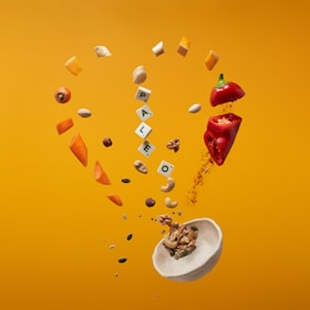 A selection of assorted nuts, fruits, seeds, and spices are artistically arranged in midair against an orange background. The word 'PALEO' is spelled out using letter tiles, surrounded by vibrant pieces of red bell pepper, carrot slices, and a scattering of seeds. A small white bowl lies below, filled with a mix of nuts being poured out.