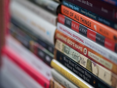 A stack of books with various titles is shown, featuring religious and spiritual themes. The spines are vertically aligned, showcasing different fonts and colors. Some titles include words like 'God' and 'Devotional,' suggesting content related to theology or religious studies. The image is focused on the books in the foreground, with the background slightly blurred.