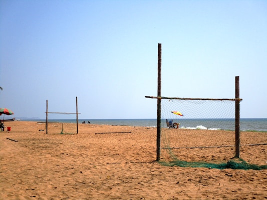 A sandy beach with makeshift wooden goalposts made from sticks and netting. In the background, a few people are gathered near the shore with a colorful beach umbrella providing shade. The sea is calm under a clear blue sky.