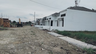A row of white houses under construction with a dirt road in front. Some construction materials and a truck are visible in the background. The sky is overcast, and patches of grass can be seen alongside the road.