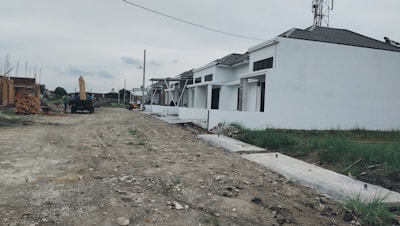A row of white houses under construction with a dirt road in front. Some construction materials and a truck are visible in the background. The sky is overcast, and patches of grass can be seen alongside the road.