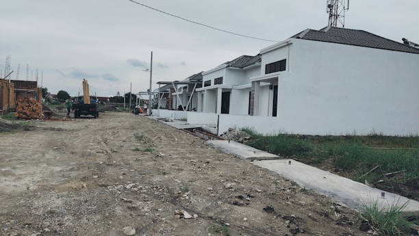 A row of white houses under construction with a dirt road in front. Some construction materials and a truck are visible in the background. The sky is overcast, and patches of grass can be seen alongside the road.