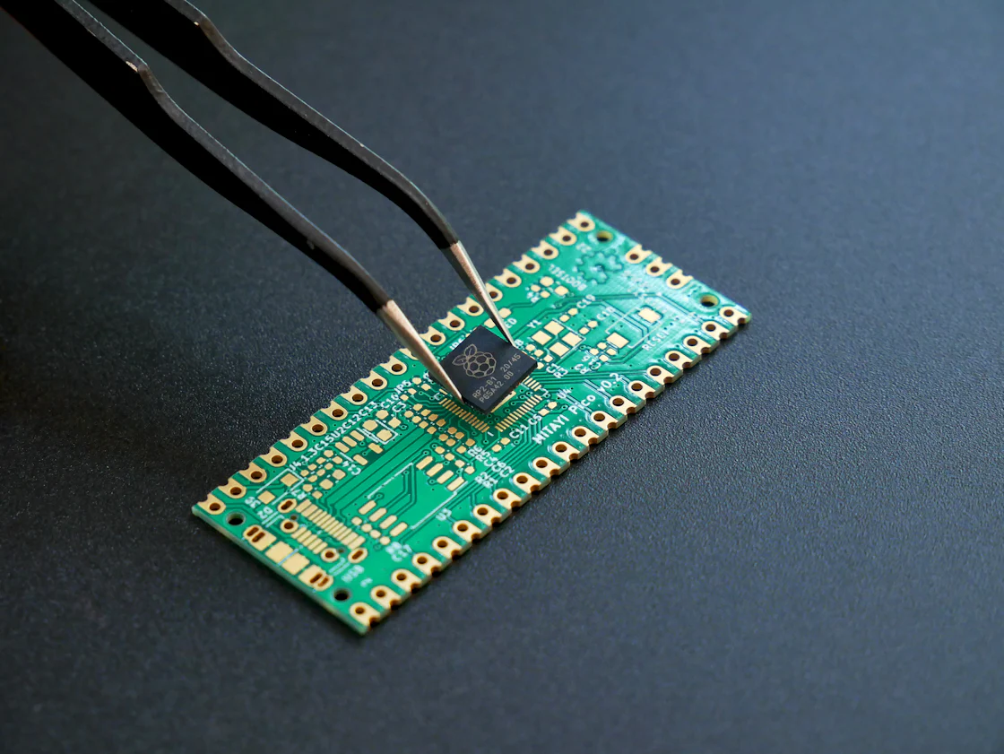 Tweezers hold a small black microchip above a green circuit board