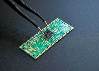 An RP2040 microcontroller held with a tweezer above a PCB. PCB was designed in KiCad and fabricated by PCBWay. Photo shot in Lumix G85.