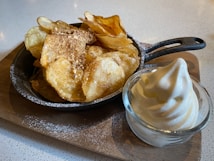 A wooden board holds a small skillet filled with crispy potato chips topped with crushed nuts, accompanied by a glass bowl of soft serve ice cream.