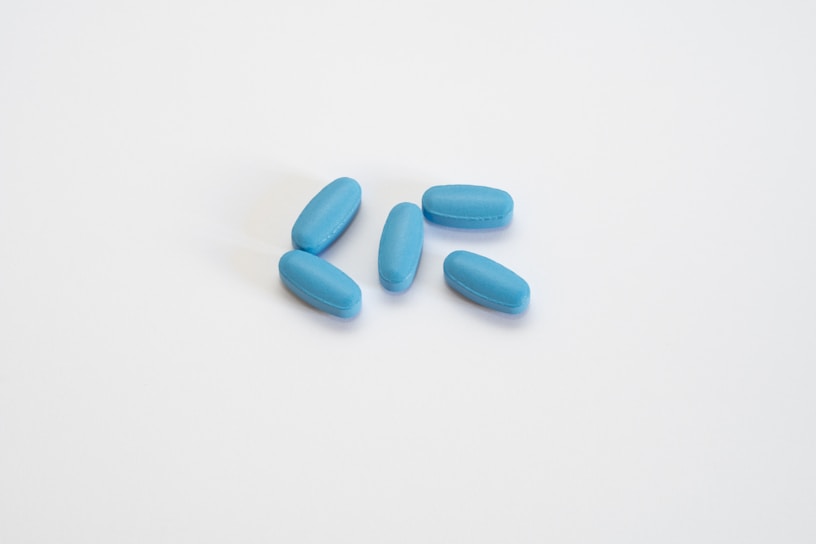 A group of six blue pills arranged on a plain white surface. The pills are oblong and evenly distributed in a slightly curved formation.