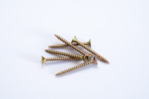 A cluster of six gold-colored screws is positioned on a clean, white surface. The screws are arranged in a slightly disordered pile, displaying their sharp threads and flat heads. Their metallic luster reflects a subtle sheen against the neutral background.