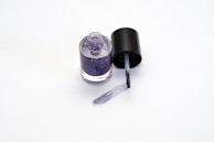 A bottle of glittery purple nail polish with the cap off and a brush resting on the surface, leaving a trail of nail polish.